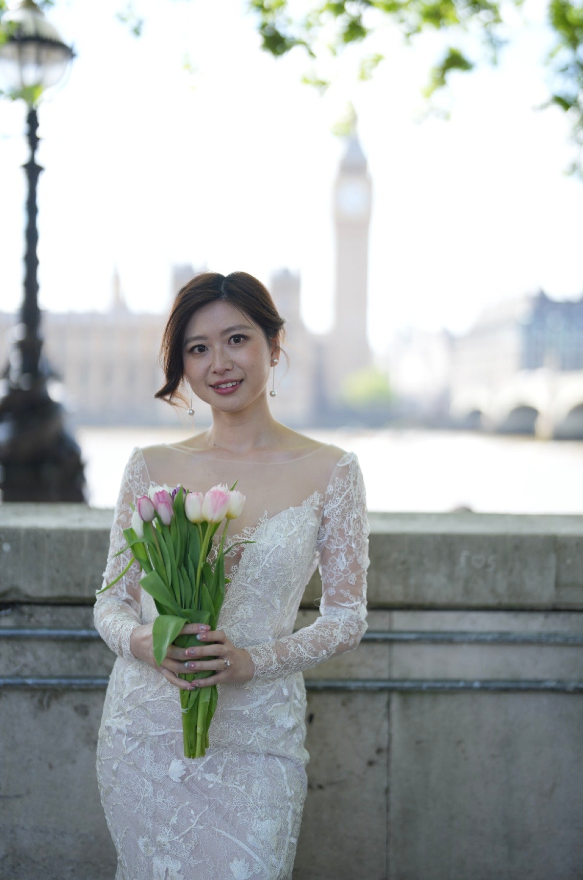 A woman wearing a white lace off-the-shoulder wedding dress stands outdoors holding a bouquet of pink and white tulips, with a blurred London landmark in the background.