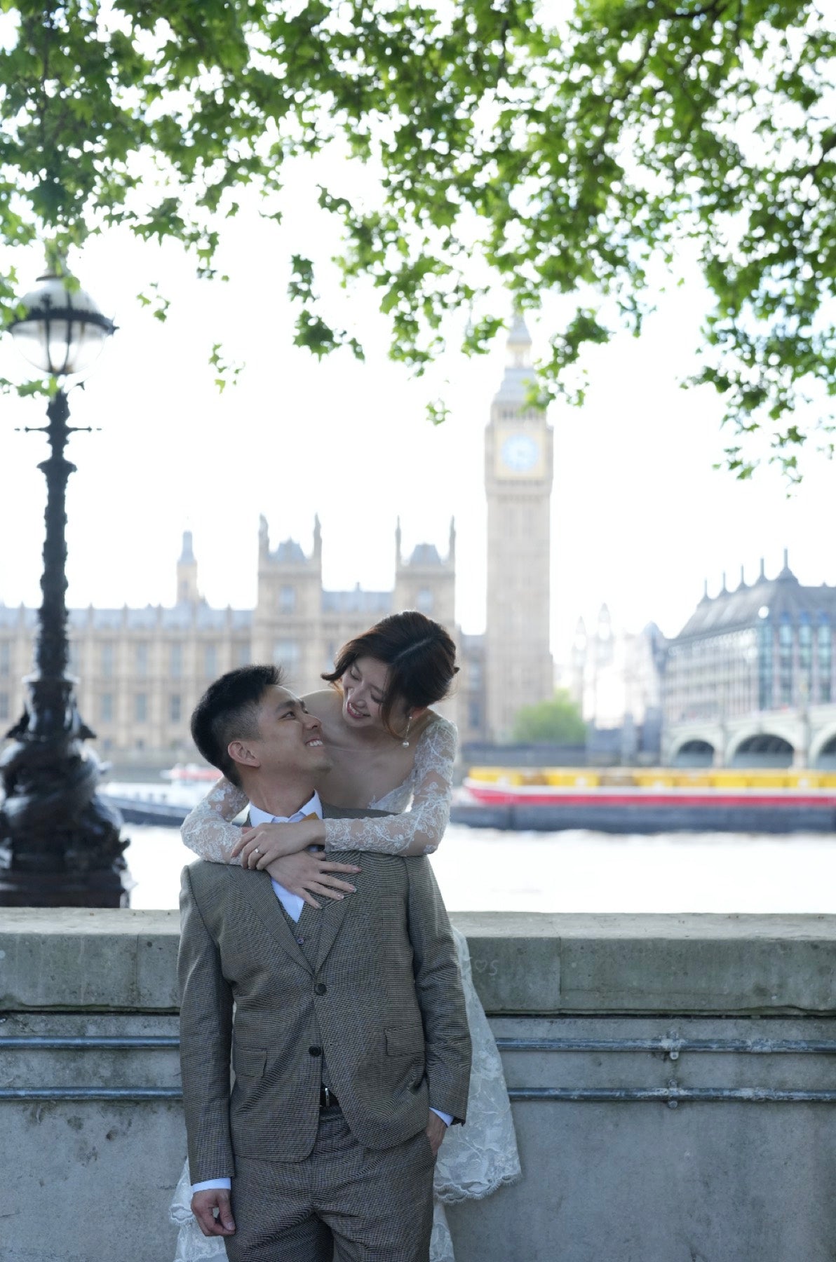 A couple embracing outdoors in front of London's Big Ben and Westminster Bridge, with leafy green branches framing the top of the photo.