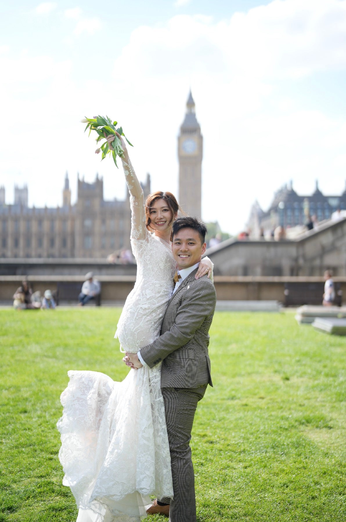 A bride in a white lace wedding gown and a groom in a gray suit pose together on a grassy lawn with Big Ben and the London Palace visible in the background.