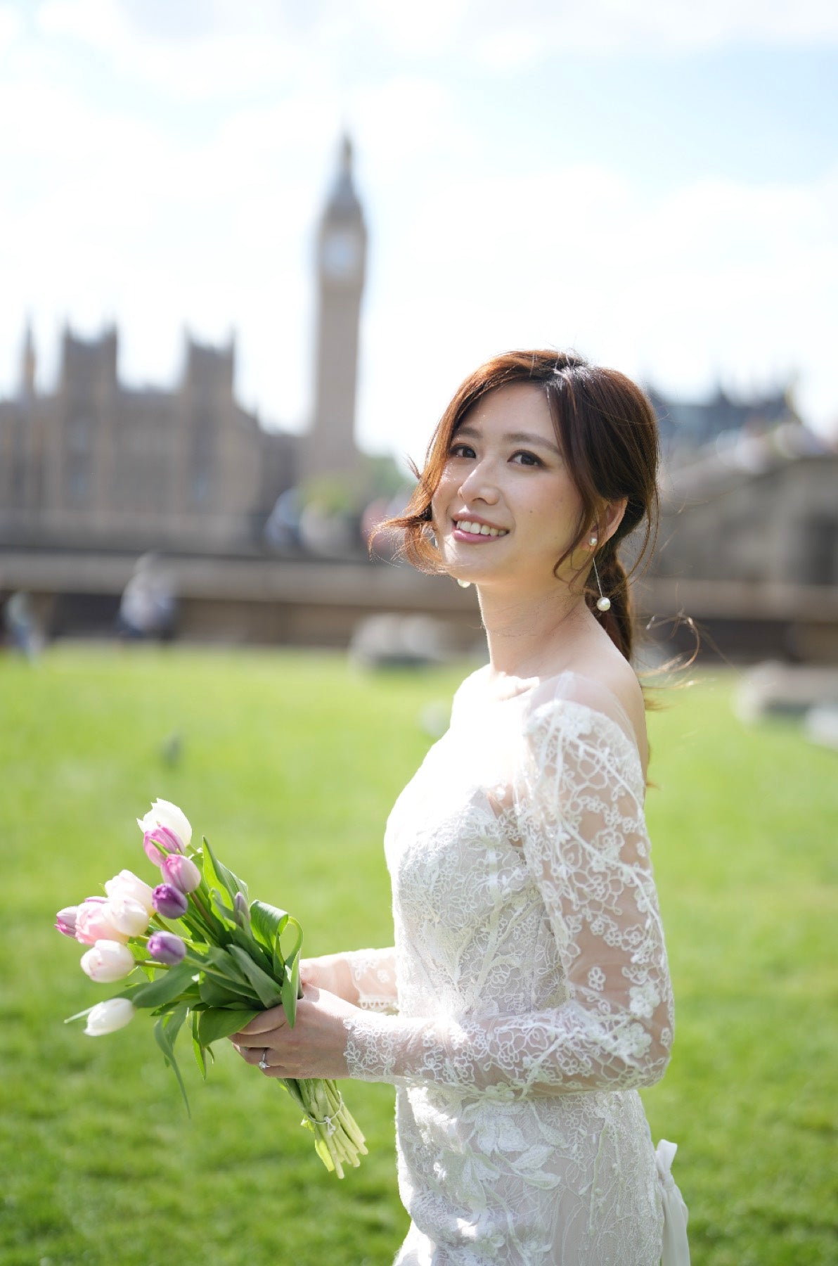 A woman in a white lace wedding dress stands outdoors on a grassy lawn holding a bouquet of pink and white tulips, with a blurred cityscape and clock tower in the background.
