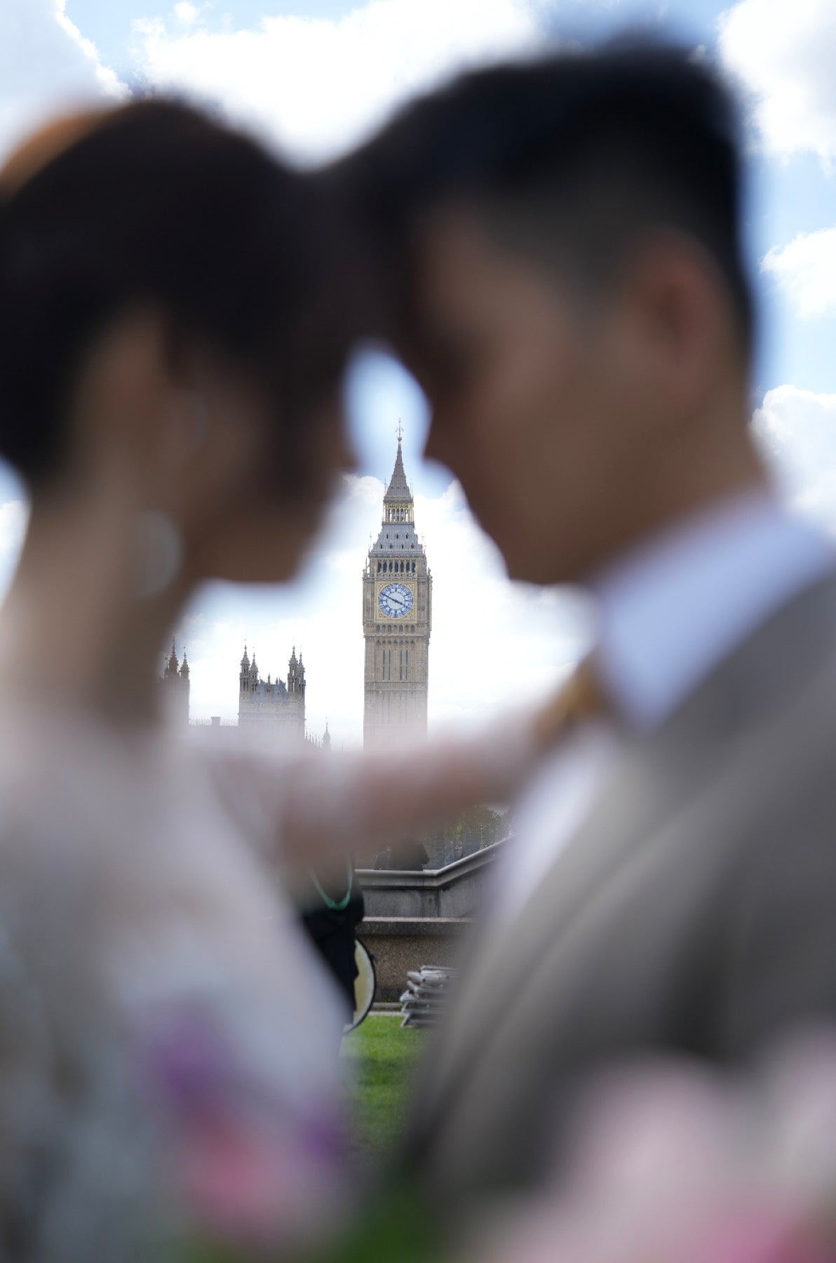 A blurred portrait of a couple in the foreground with Big Ben and Elizabeth Tower visible in the background under a cloudy sky.