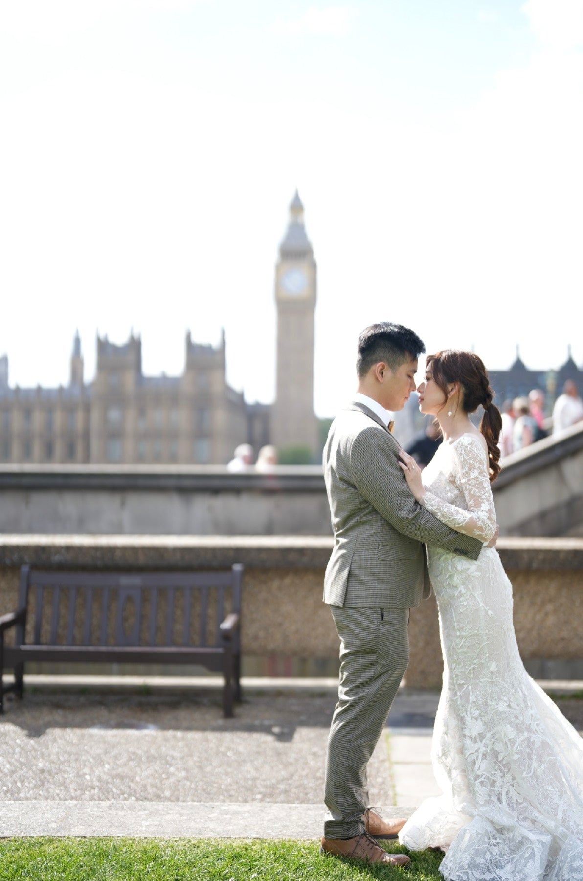 A bride in a white lace wedding gown and a groom in a gray suit embrace and kiss outdoors, with Big Ben and the Houses of Parliament blurred in the background.