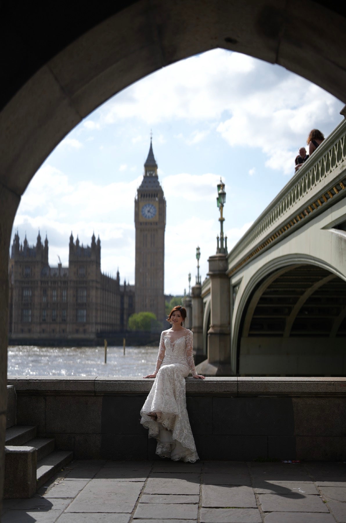 A woman in a white wedding gown sits on a stone ledge in front of a large stone archway, with Big Ben and Westminster Bridge in London in the background.