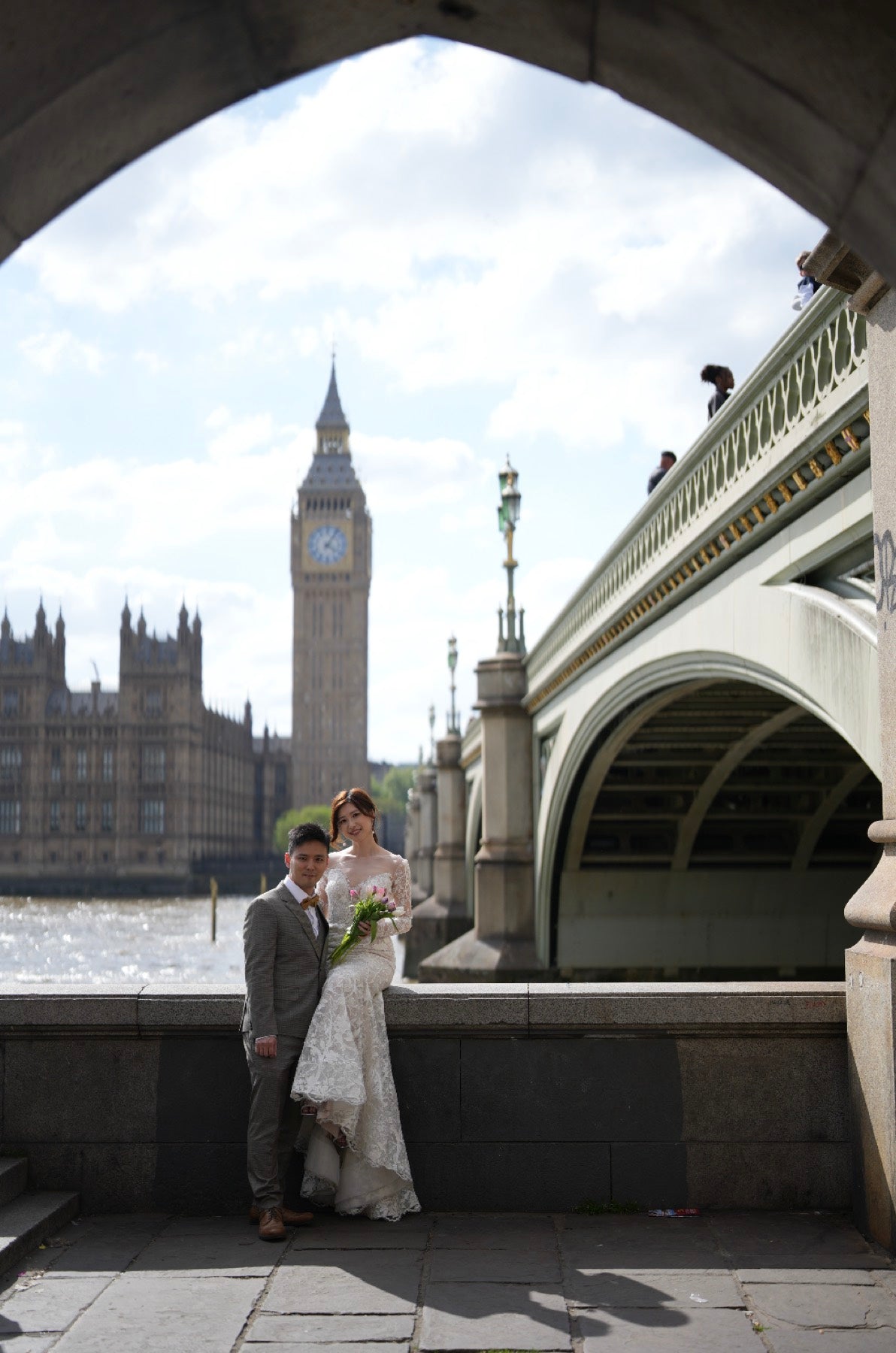 A bride in a white wedding gown and a groom in a gray suit pose together under a stone archway, with Big Ben and Westminster Bridge in London in the background.