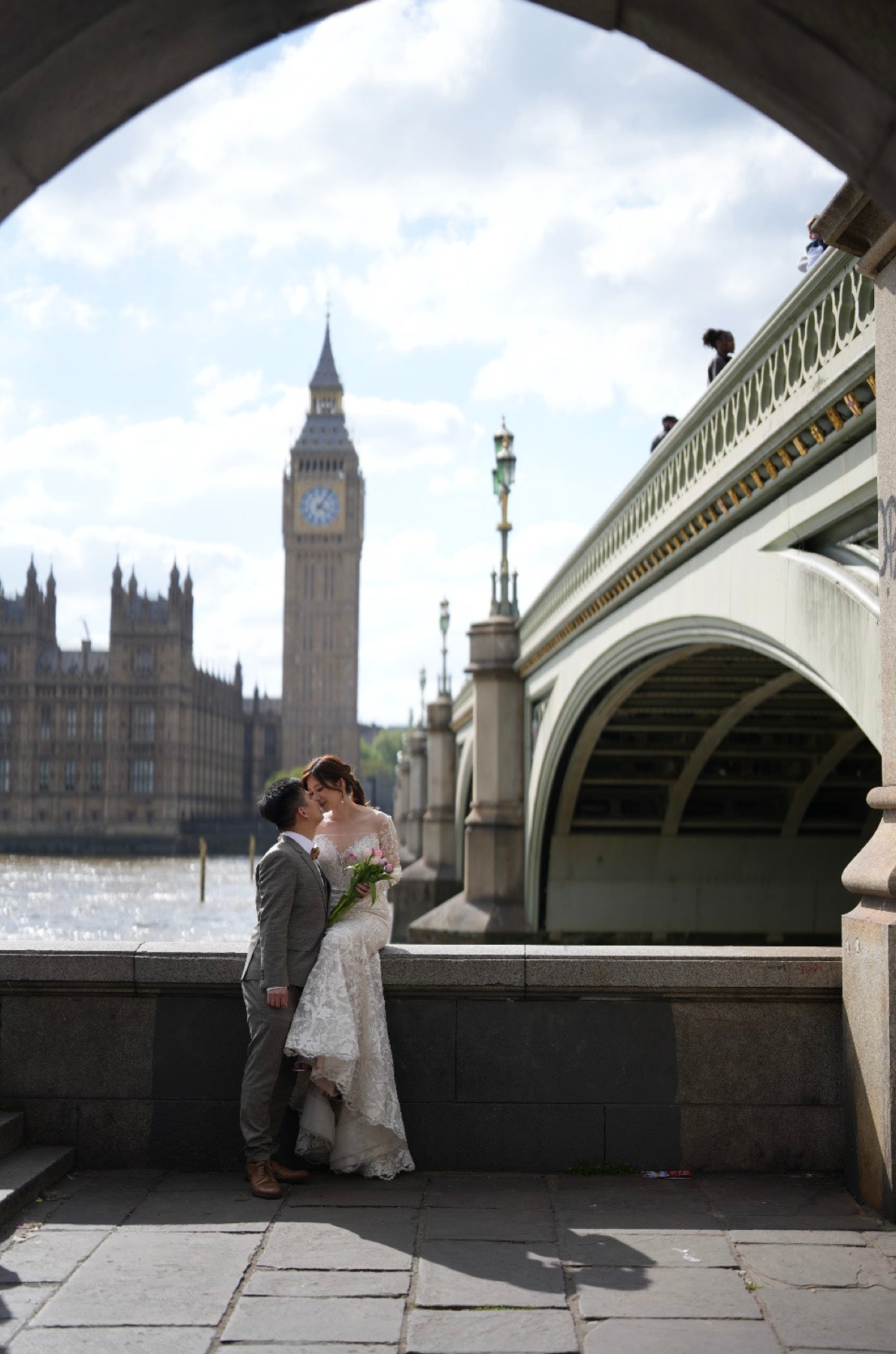 A bride in a white lace wedding gown and a groom in a gray suit kiss under a stone archway, with Big Ben and the Houses of Parliament in London and Westminster Bridge in the background.