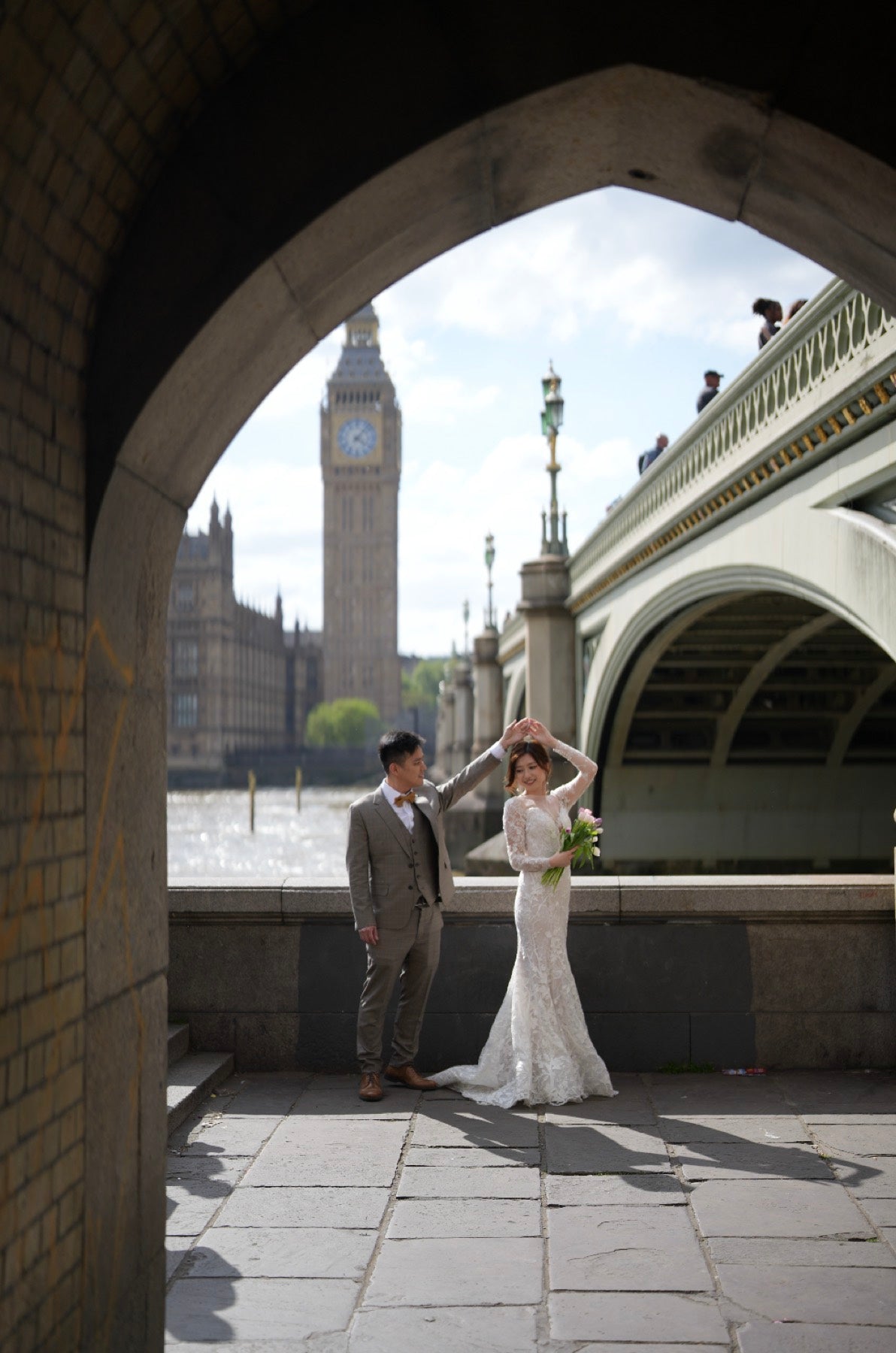 A bride in a white lace wedding gown and a groom in a gray suit pose under a stone archway, with Big Ben and Westminster Bridge in London in the background.