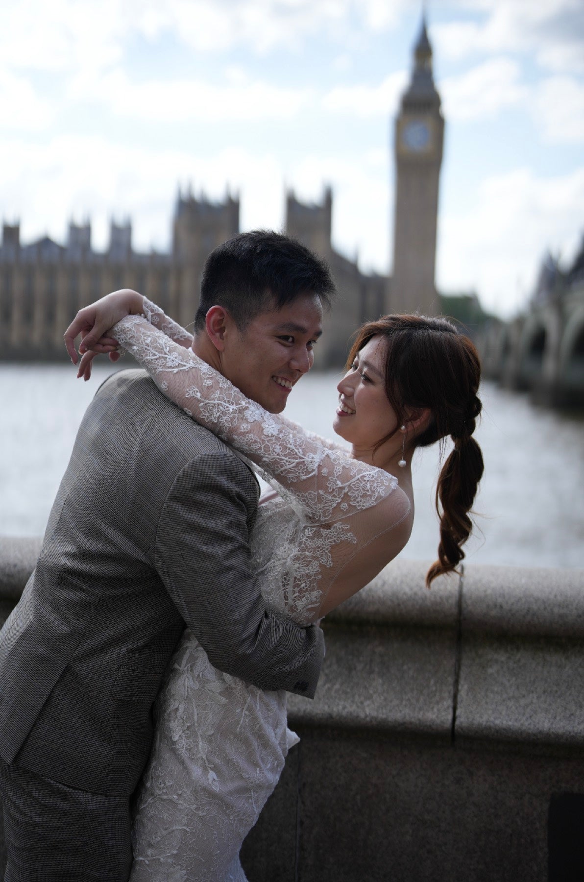 A couple embracing on a bridge with London's Big Ben and Houses of Parliament in the background. The man is wearing a gray suit, and the woman is wearing a white lace wedding dress.