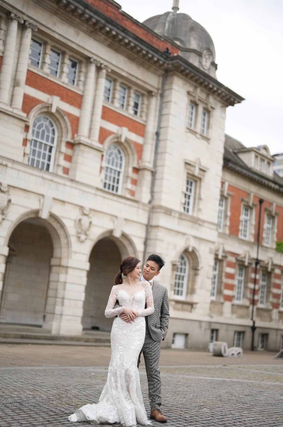 A bride in a white lace wedding gown and a groom in a gray suit standing together in front of a large historic stone building with red brick accents.