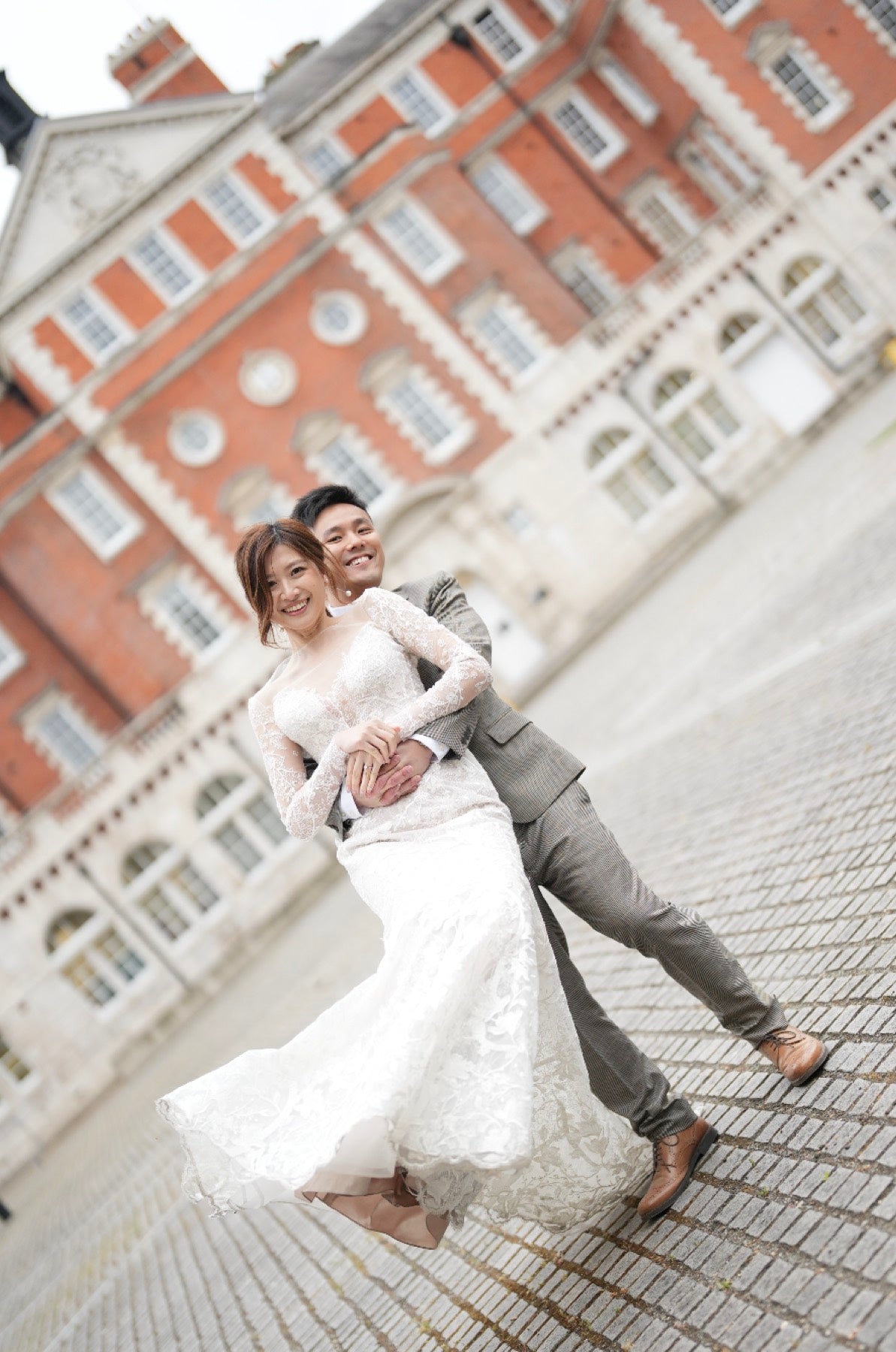 A bride in a white lace wedding gown and a groom in a gray suit pose together on a cobblestone street in front of a large red brick building.