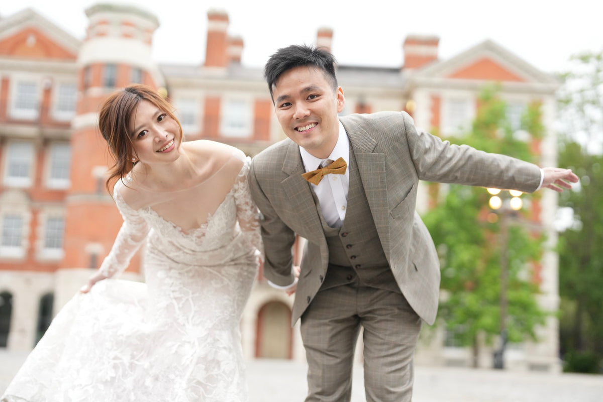 A smiling bride in a white lace wedding gown and a groom in a gray three-piece suit with a brown bow tie pose playfully in front of a red brick college-style building.