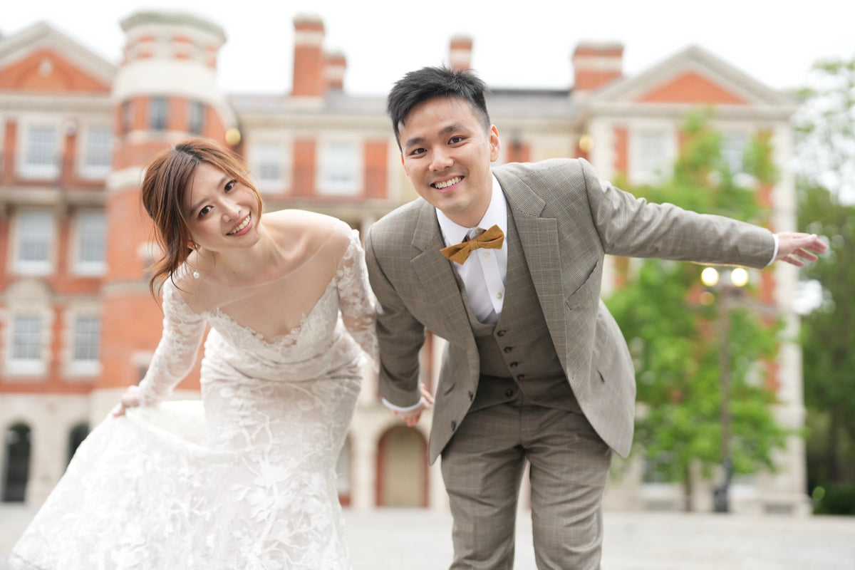 A smiling couple posing outdoors in front of a large red brick building. The woman wears an off-the-shoulder white lace wedding gown, and the man wears a gray three-piece suit with a brown bow tie.