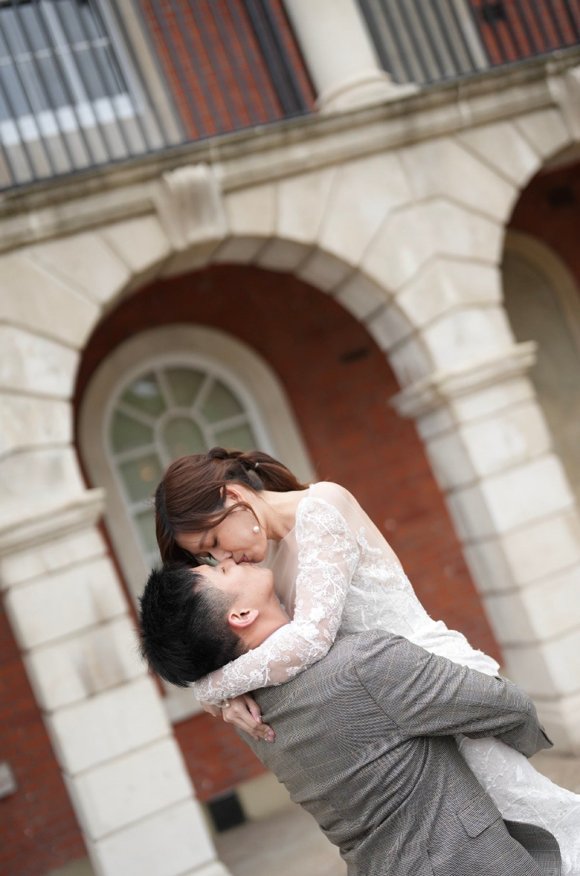 A couple embracing and kissing outdoors in front of a stone building with red brick arches. The woman wears a white lace dress, and the man wears a gray suit.