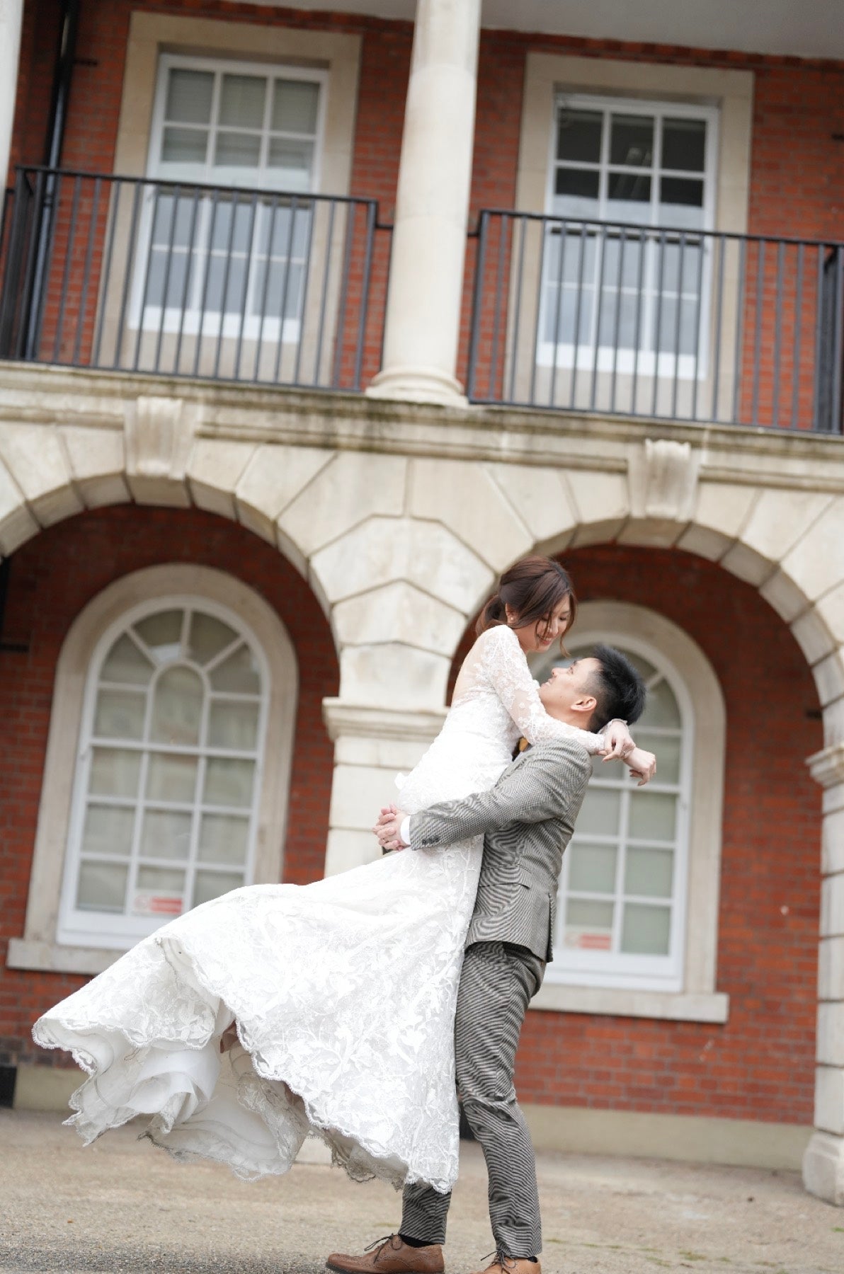A bride in a white lace wedding gown and a groom in a gray suit are captured mid-spin as she winds her dress around him, set against the brick and stone facade of a classic building.