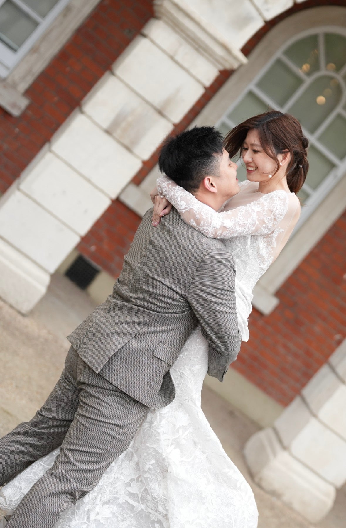 A bride in a white lace wedding gown and a groom in a gray suit embrace and kiss outdoors in front of a red brick building with white stone columns.