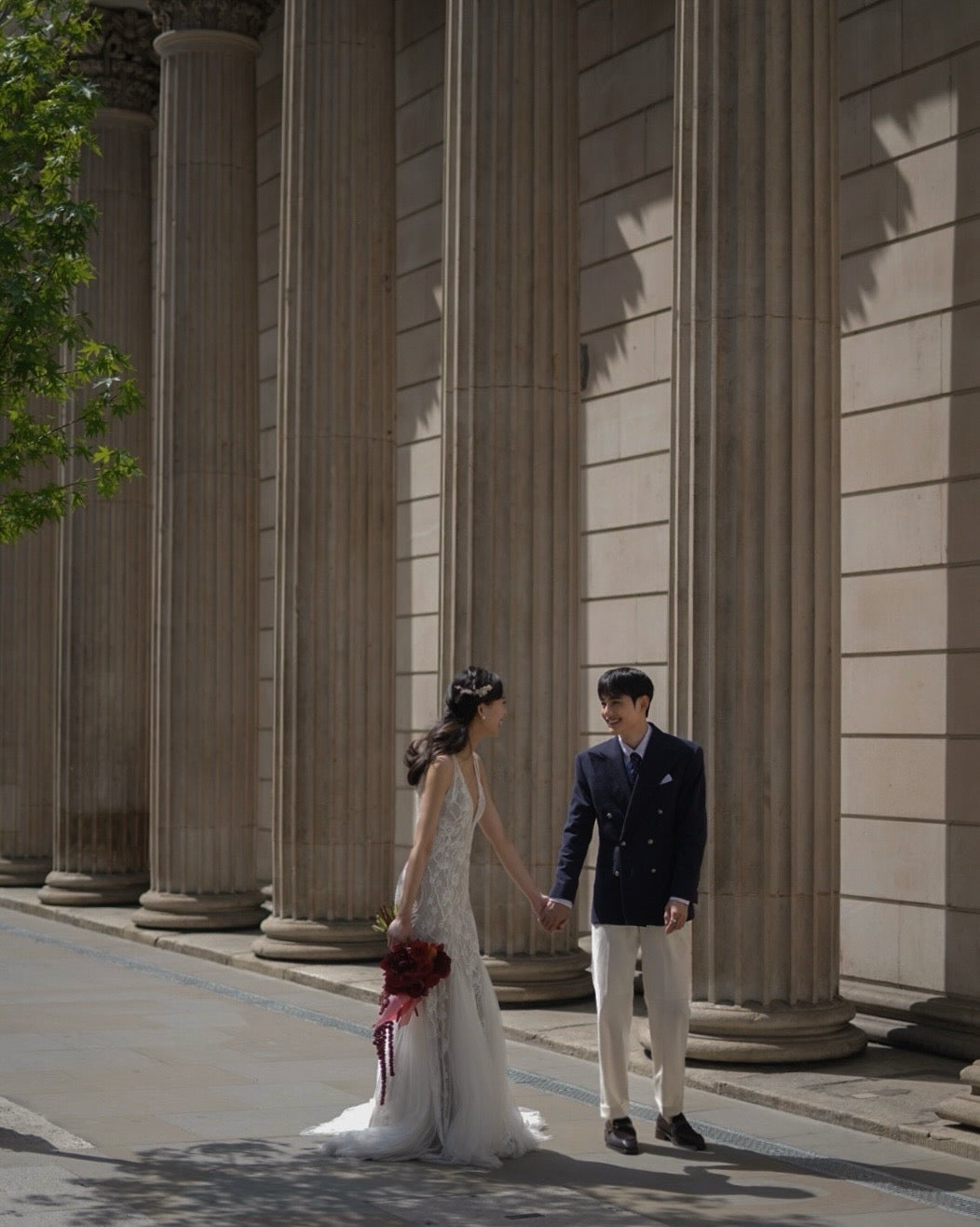 A bride in a sleeveless white lace wedding gown holding hands with a groom wearing a navy blazer, cream trousers, and dark loafers, standing in front of a grand classical stone colonnade.