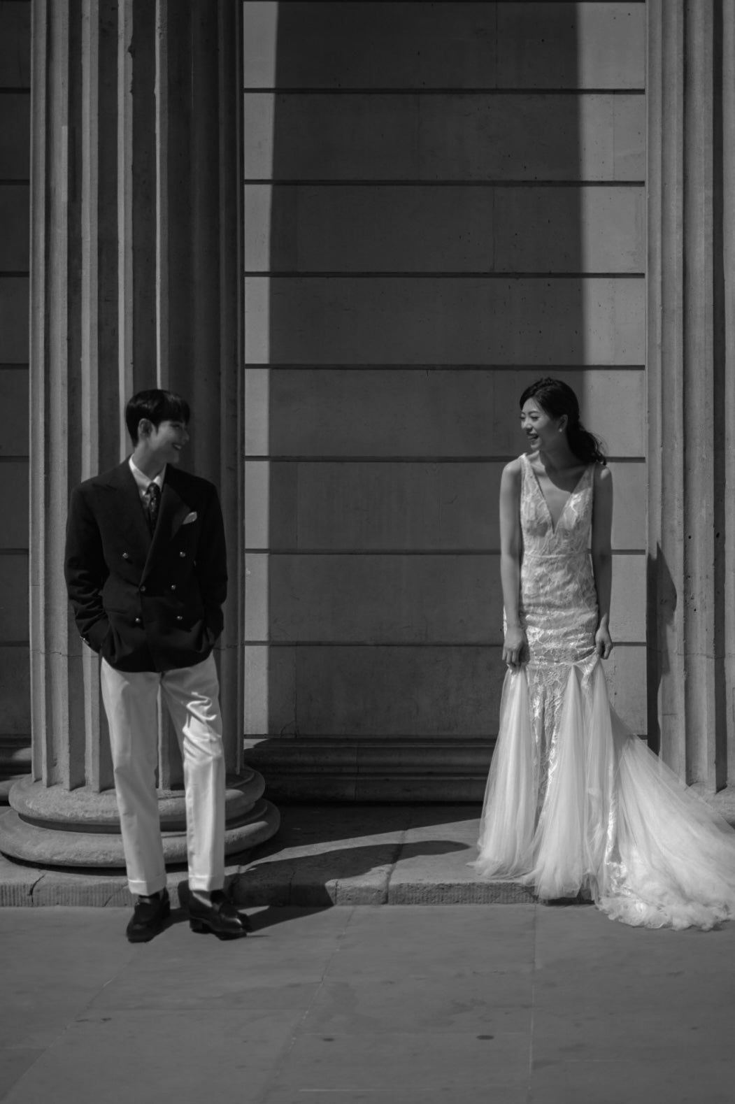 A black and white photograph of a bride in a sleeveless lace wedding gown and a groom in a dark blazer and light trousers standing in front of classical stone columns.