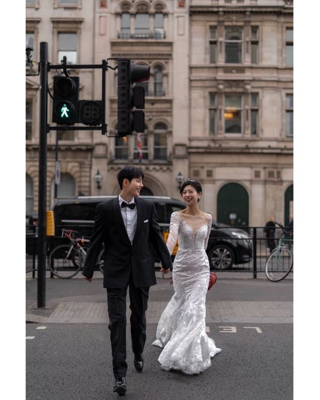 A bride in a white lace wedding gown and a groom in a black tuxedo walk hand-in-hand across a city street in front of a historic stone building.