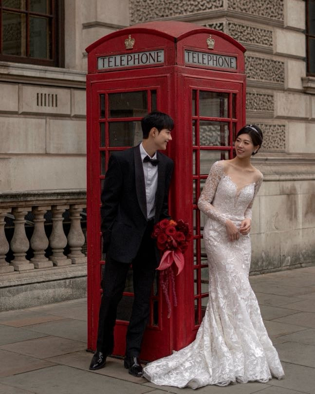 A bride in a white lace mermaid wedding gown and a groom in a black tuxedo stand beside a classic red London telephone booth on a city street.