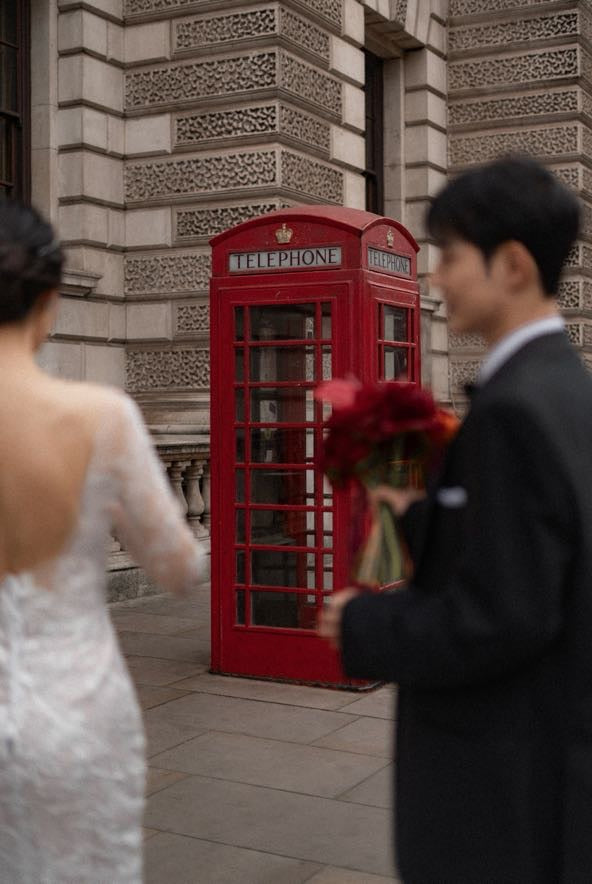 A red classic London telephone booth stands on a stone sidewalk in front of a building facade. A bride in a white wedding dress is seen from behind on the left, and a groom in a dark suit holding a bouquet of red flowers is blurred on the right.
