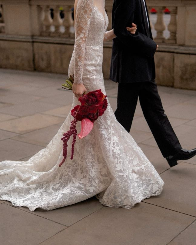 A bride in a long-sleeved white lace wedding gown walking outdoors beside a man in a black suit, holding a red floral bouquet.