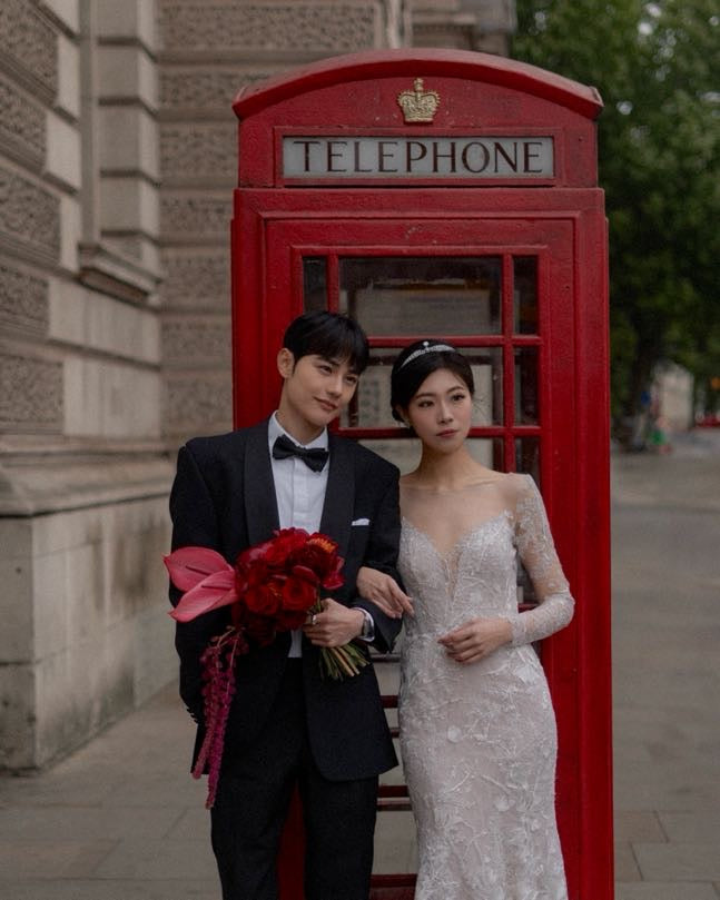 A bride in a white lace wedding gown and a groom in a black tuxedo with a bow tie stand in front of a classic red British telephone booth, holding a bouquet of red roses.