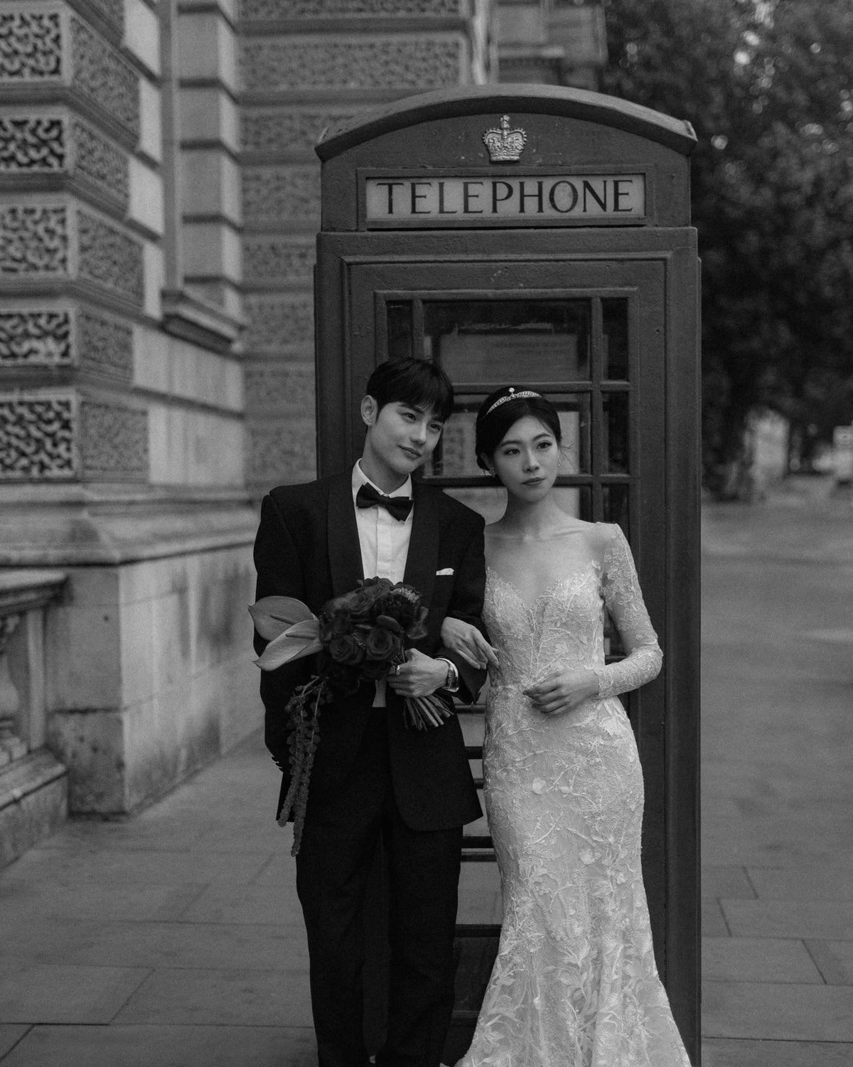 A black and white photograph of a bride and groom standing beside a classic British telephone booth in front of a stone building. The groom wears a black tuxedo with a bow tie and holds a bouquet, while the bride wears a fitted lace wedding gown.