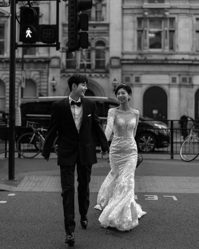 A black and white photograph of a bride and groom walking hand-in-hand across a city street. The groom is wearing a classic black tuxedo with a white shirt and bow tie, and the bride is wearing a fitted lace wedding gown.
