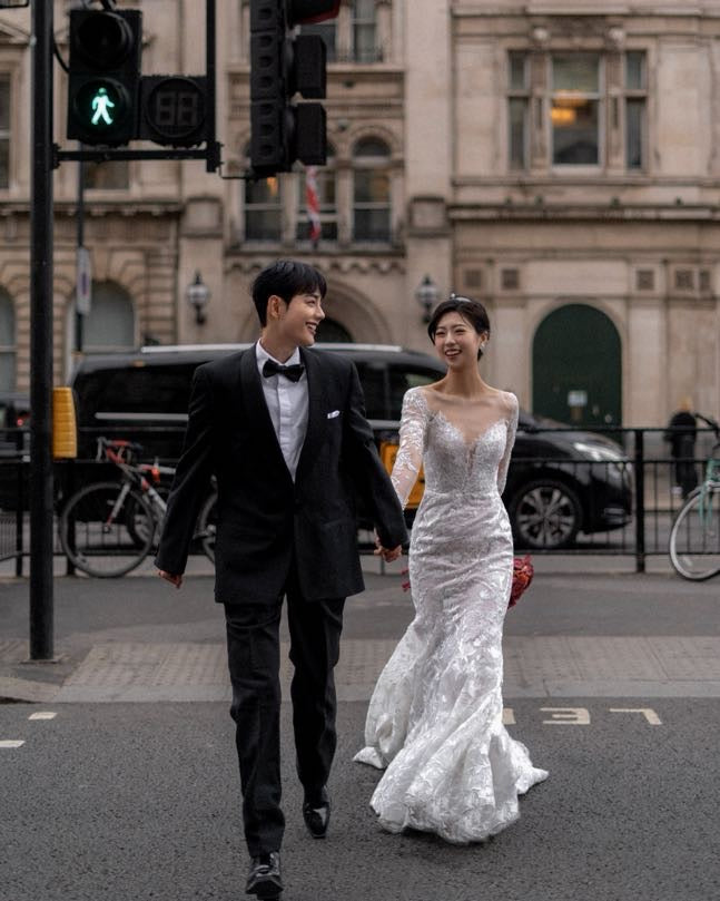 A bride in a fitted white lace wedding gown and a groom in a black tuxedo walk hand-in-hand across a city street in front of a historic stone building.