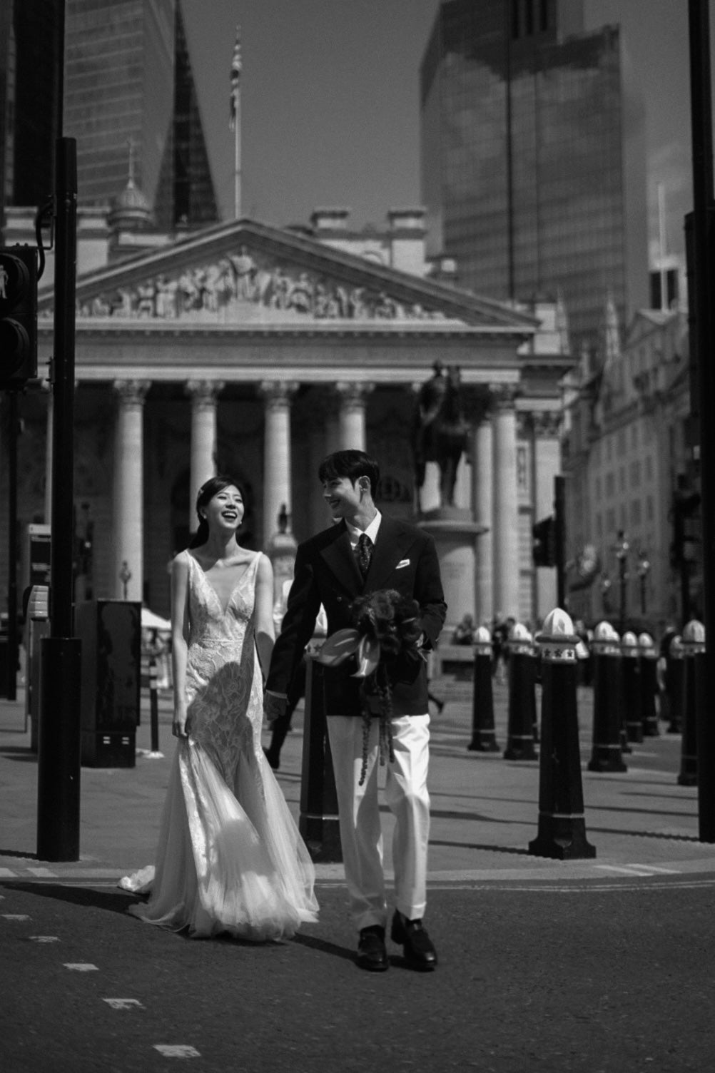 A black and white photograph of a bride in a sleeveless lace wedding gown and a groom in a dark suit and white trousers walking hand in hand on a city street, with a grand neoclassical building in the background.