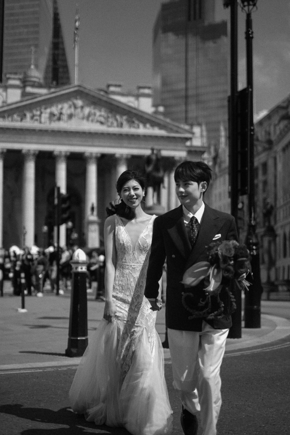 A black and white photograph of a bride and groom walking hand in hand on a city street. The bride is wearing a sleeveless lace wedding gown, and the groom is in a dark suit with white trousers.