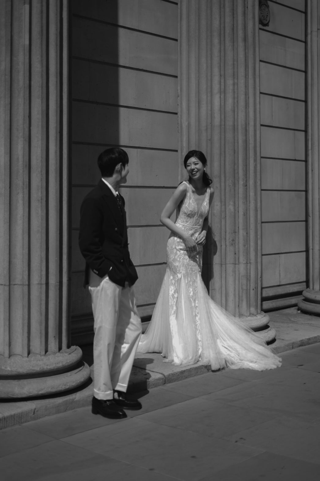 Black and white photograph of a bride and groom standing in front of classical stone columns. The bride is wearing a sleeveless lace mermaid-style wedding gown with a long train, and the groom is wearing a dark jacket, white trousers, and dark shoes.