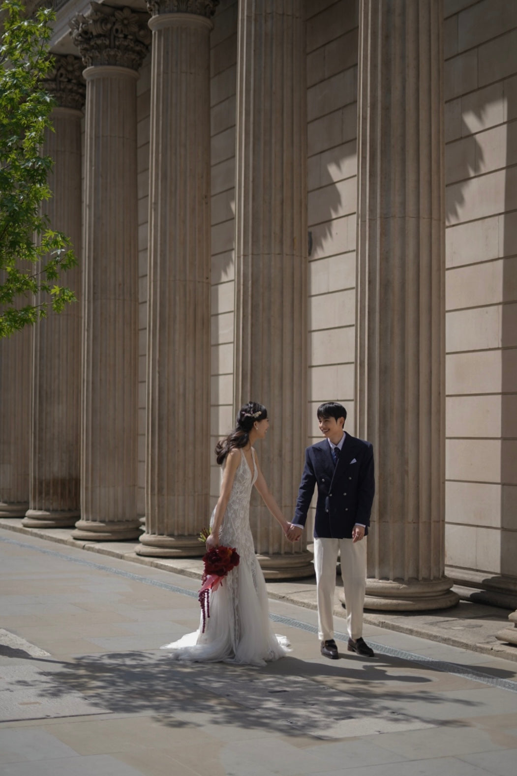 A bride in a sleeveless white wedding gown holding hands with a groom wearing a dark navy double-breasted suit, cream trousers, and black dress shoes, standing in front of a grand building with large stone columns.