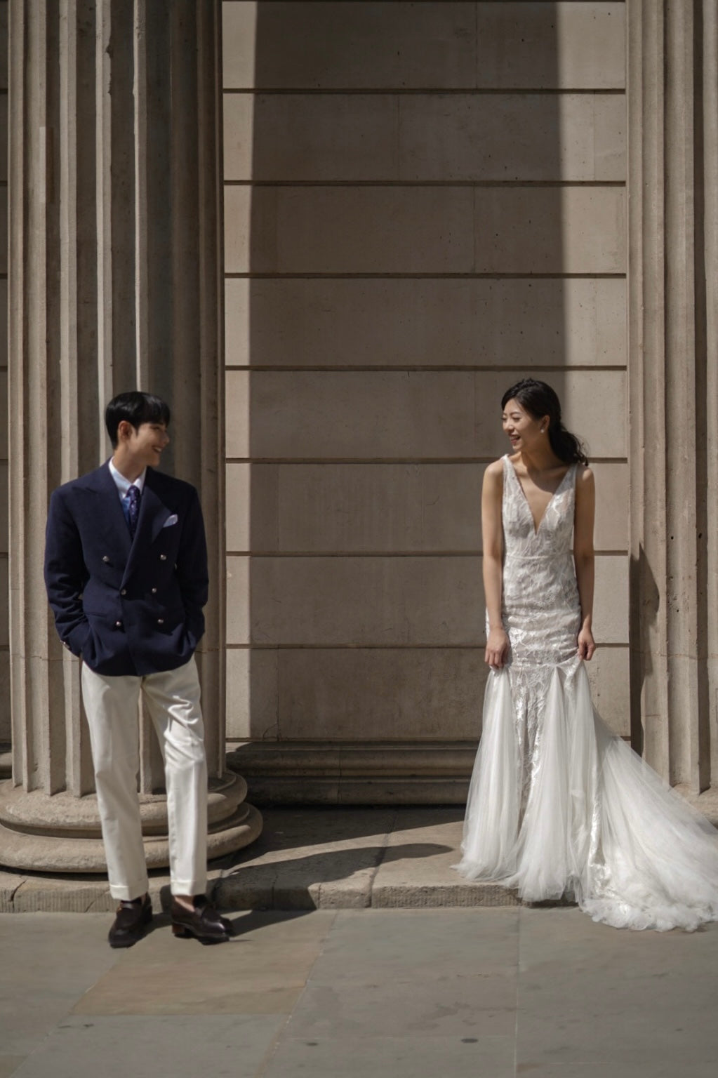 A bride in a white lace mermaid wedding gown and a groom in a navy double-breasted suit and cream trousers pose together outdoors against a stone building with columns.