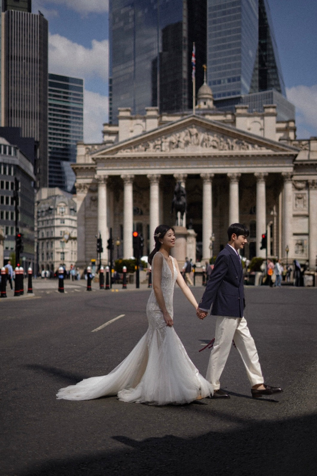 A bride in a white sleeveless mermaid wedding gown and a groom in a navy blazer and cream trousers walk hand-in-hand across a city street in front of a grand neoclassical building and modern skyscrapers.