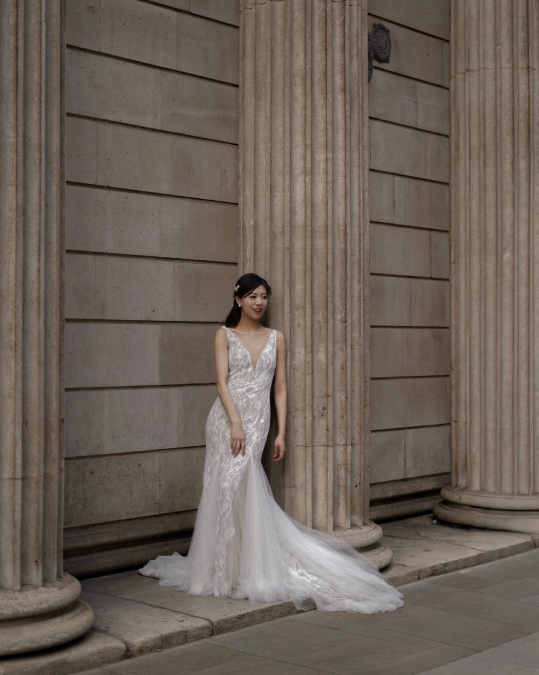 A woman wearing a sleeveless white lace wedding gown with a deep V-neckline and a long flowing train, standing in front of large stone columns.