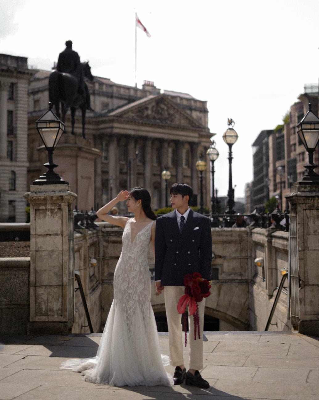 A bride in a sleeveless white lace wedding gown and a groom in a dark double-breasted suit with cream trousers stand together on a stone bridge in front of a classical building and equestrian statue.