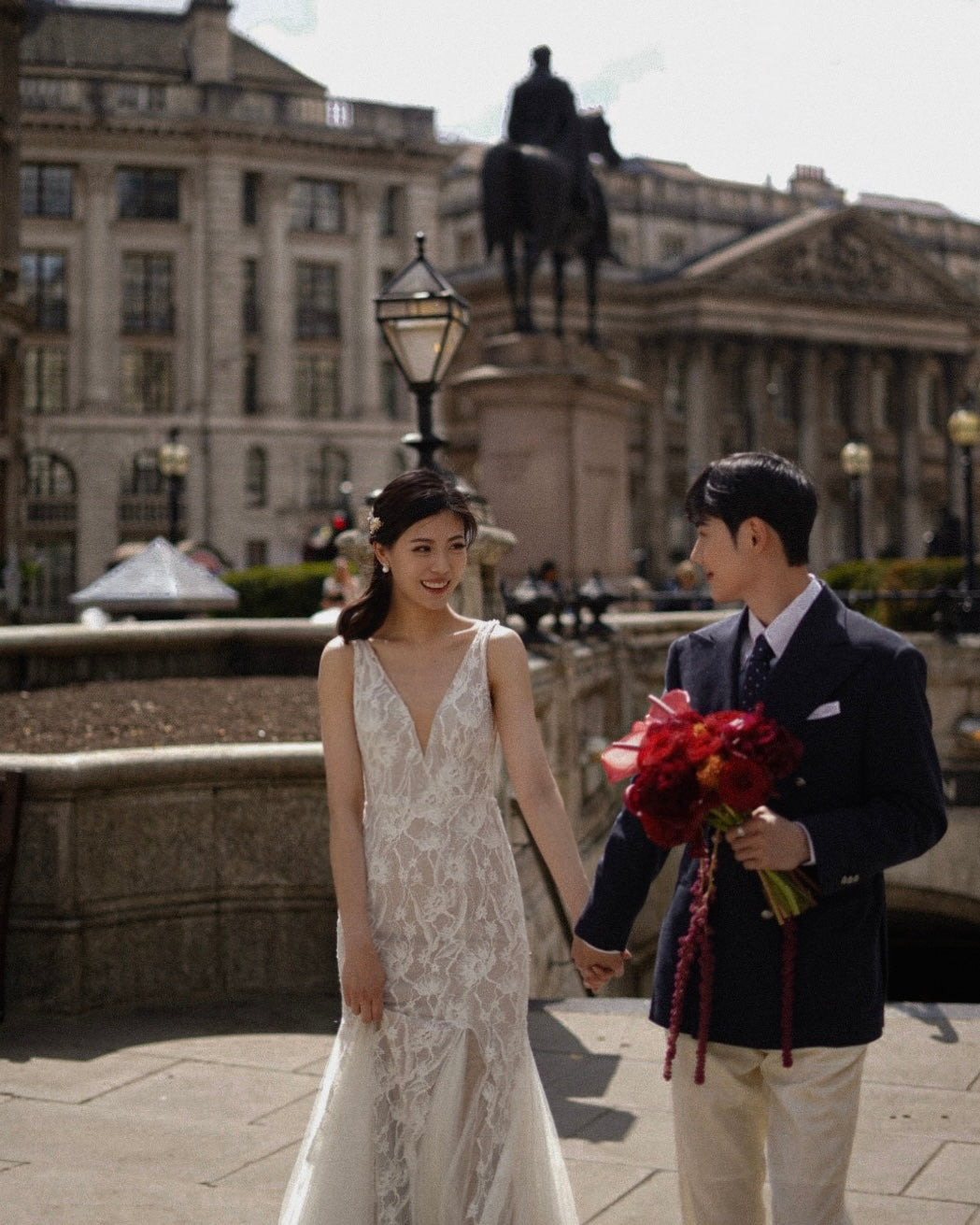 A bride in a sleeveless white lace wedding gown and a groom in a navy suit holding a bouquet of red flowers, walking hand-in-hand in an outdoor urban setting with a historic building and equestrian statue in the background.