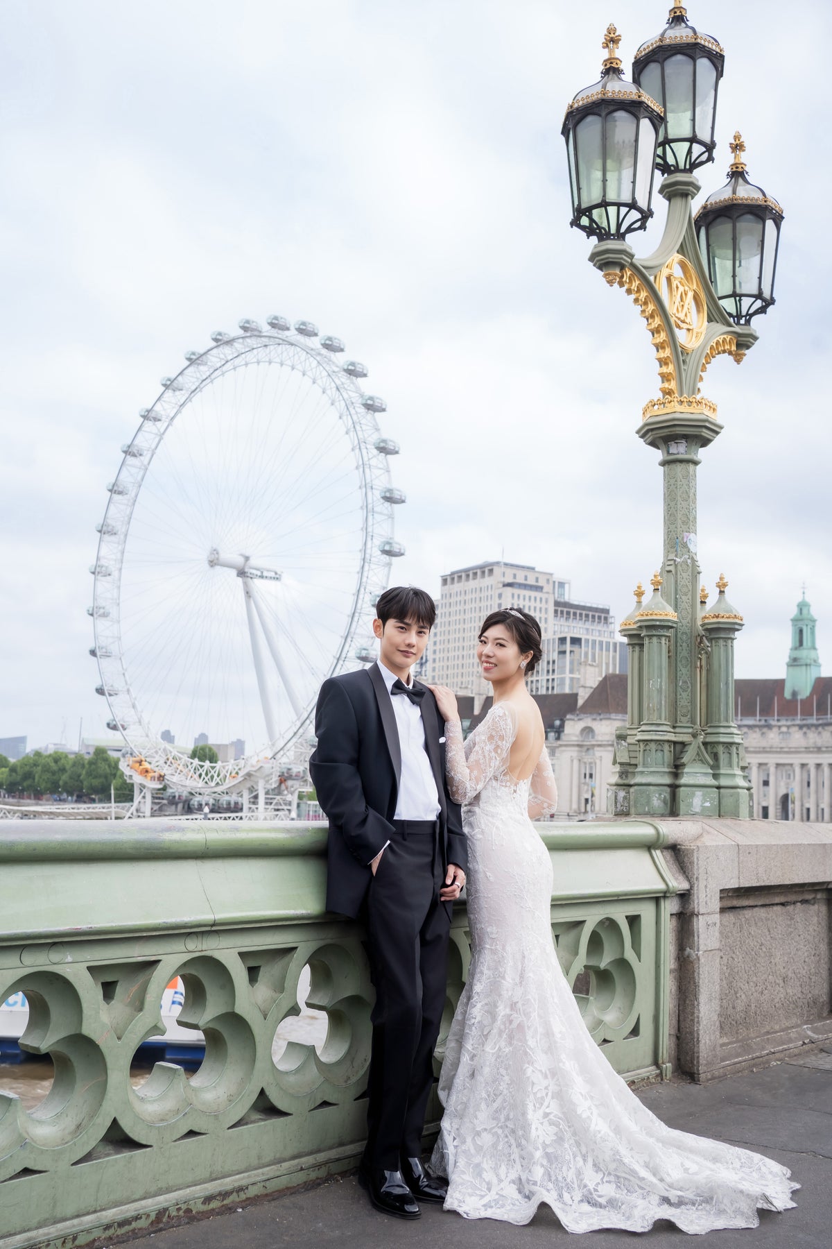 A bride in a white lace wedding gown and a groom in a black tuxedo stand together on a bridge, with the London Eye and a decorative lamp post in the background.