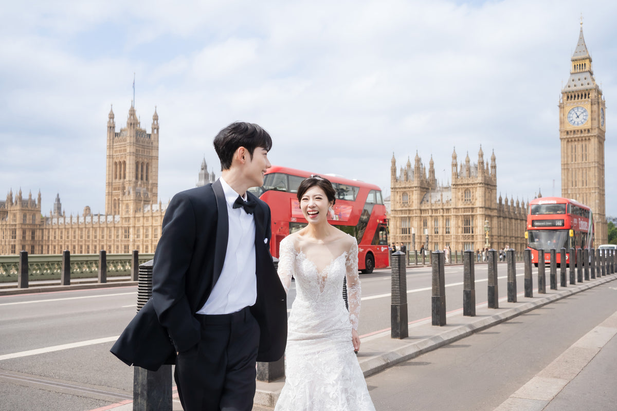 A bride in a white lace wedding gown and a groom in a black tuxedo with a bow tie walk hand in hand on London Bridge, with Big Ben and the Houses of Parliament in the background.