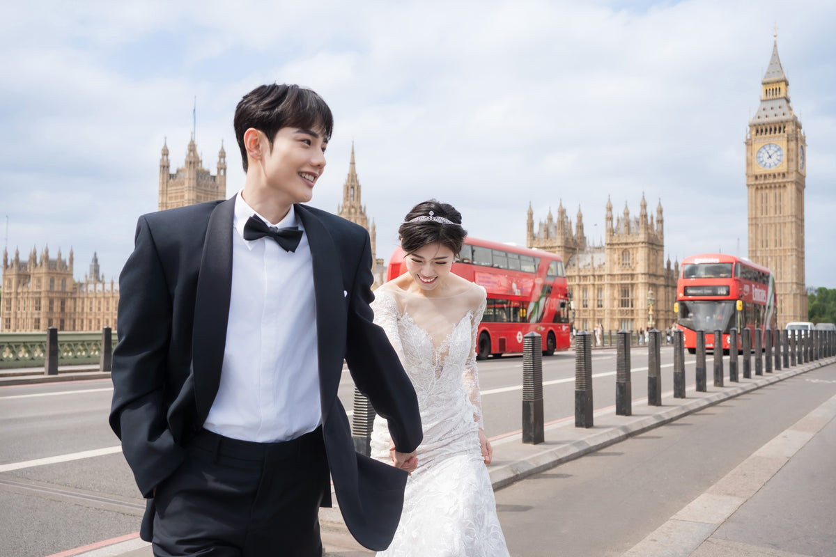 A smiling man in a black tuxedo and a woman in a white wedding gown walk hand in hand along Westminster Bridge in London, with Big Ben and the Houses of Parliament in the background, alongside red double-decker buses.