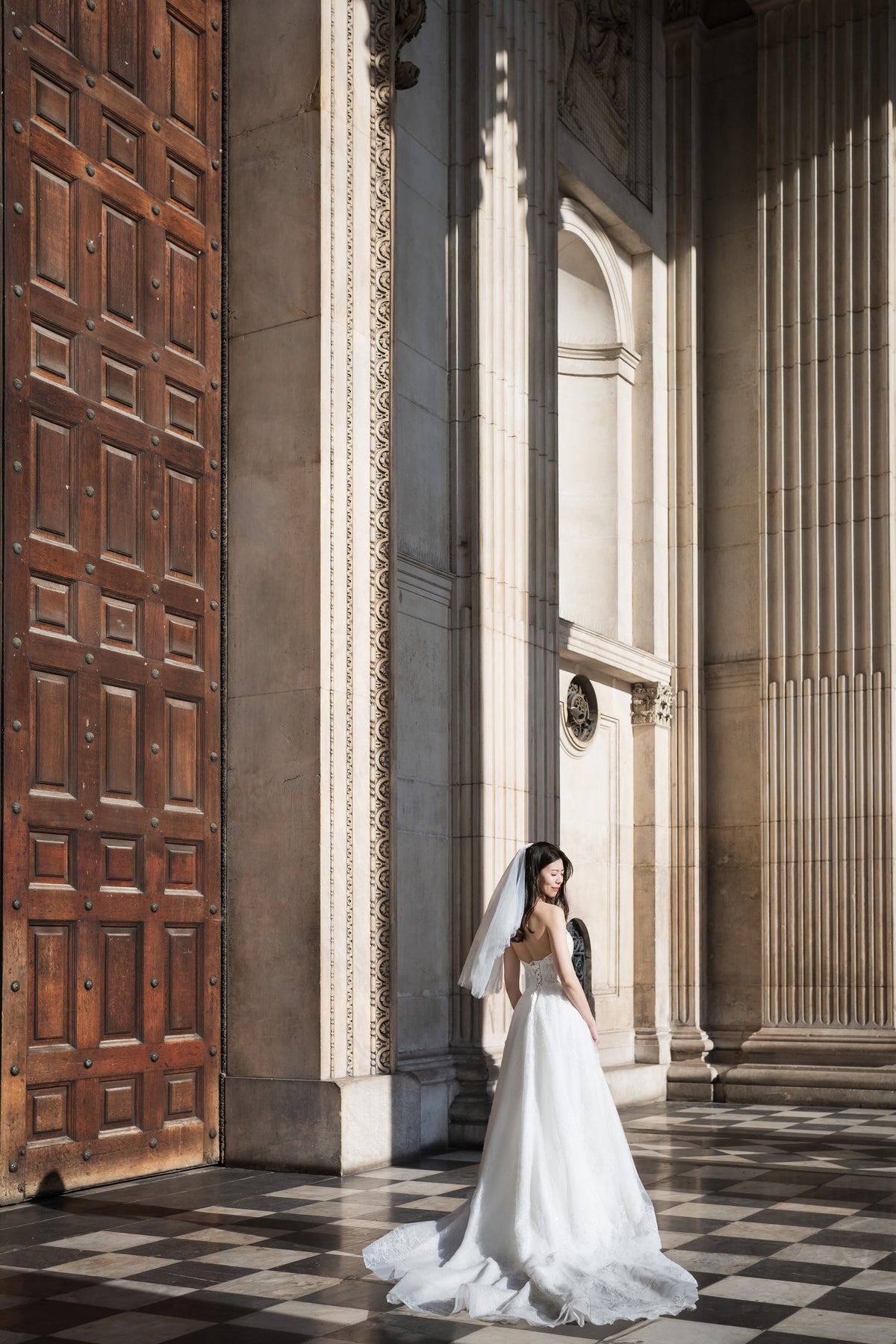 A woman in a white wedding gown and veil stands in a grand stone corridor with a large wooden door on the left, sunlight casting strong shadows across the floor.