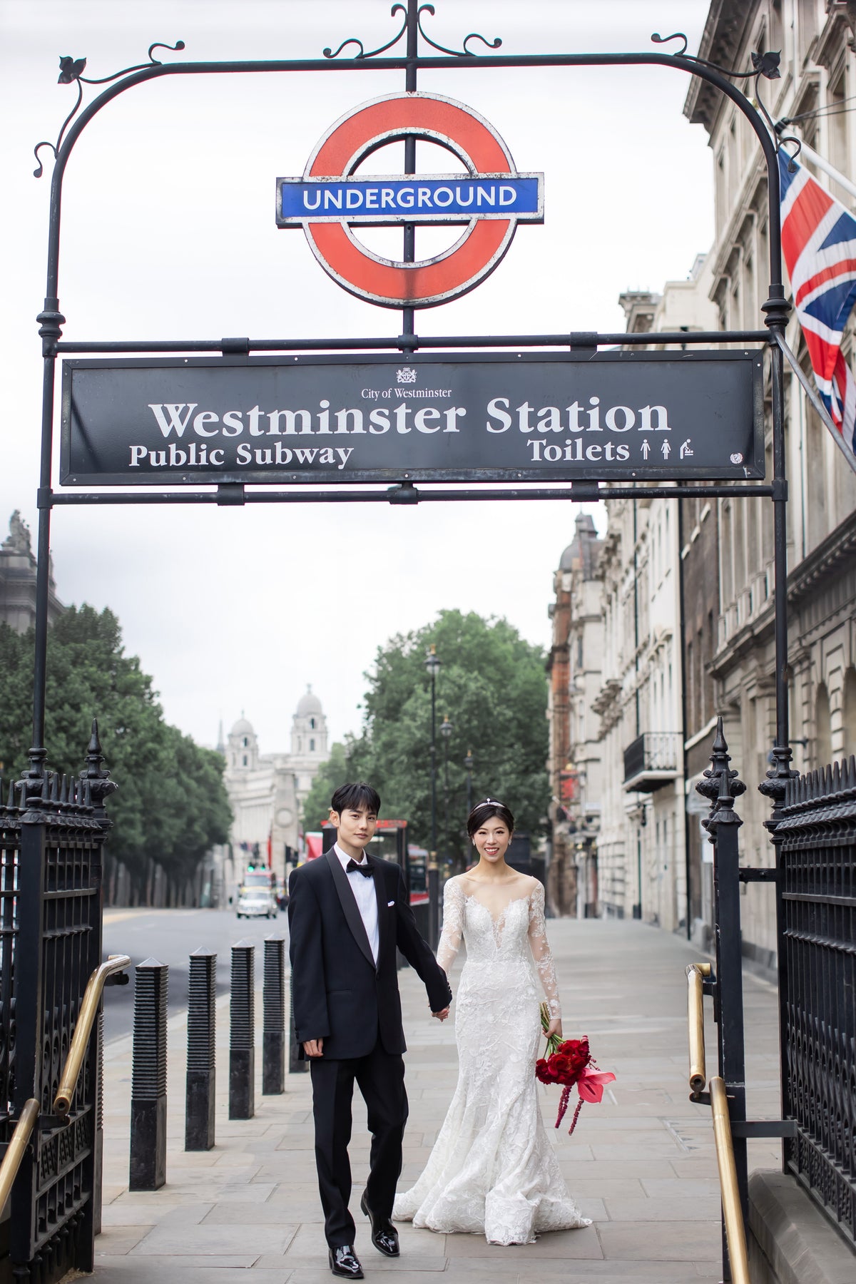 A bride in a white lace wedding gown and a groom in a black tuxedo walk hand-in-hand through the entrance arch of Westminster Station Underground station in London, with the iconic red and blue roundel sign visible above them.