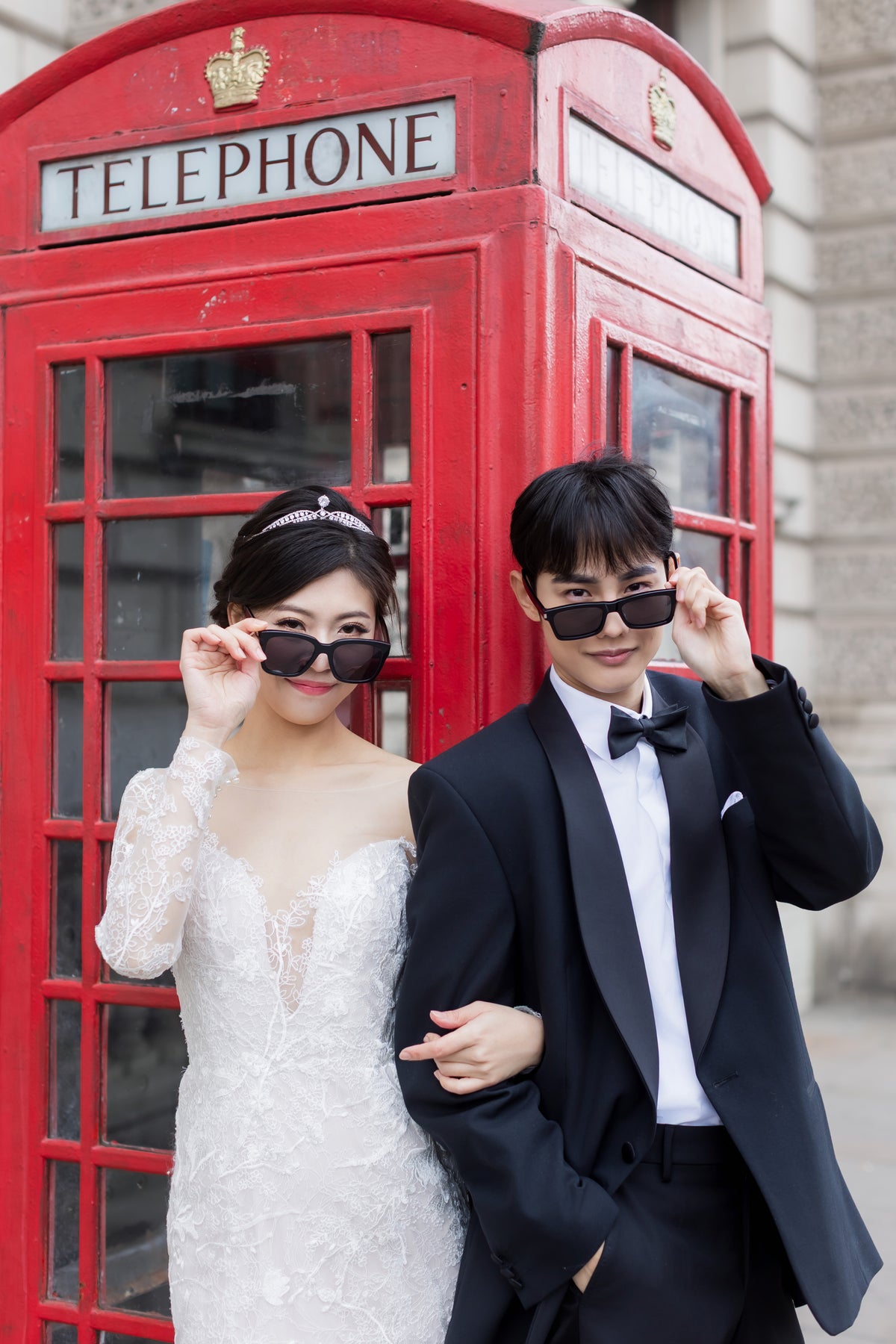 A bride in a white lace wedding dress and a groom in a black tuxedo with a bow tie pose in front of a classic red telephone booth, both wearing black sunglasses.