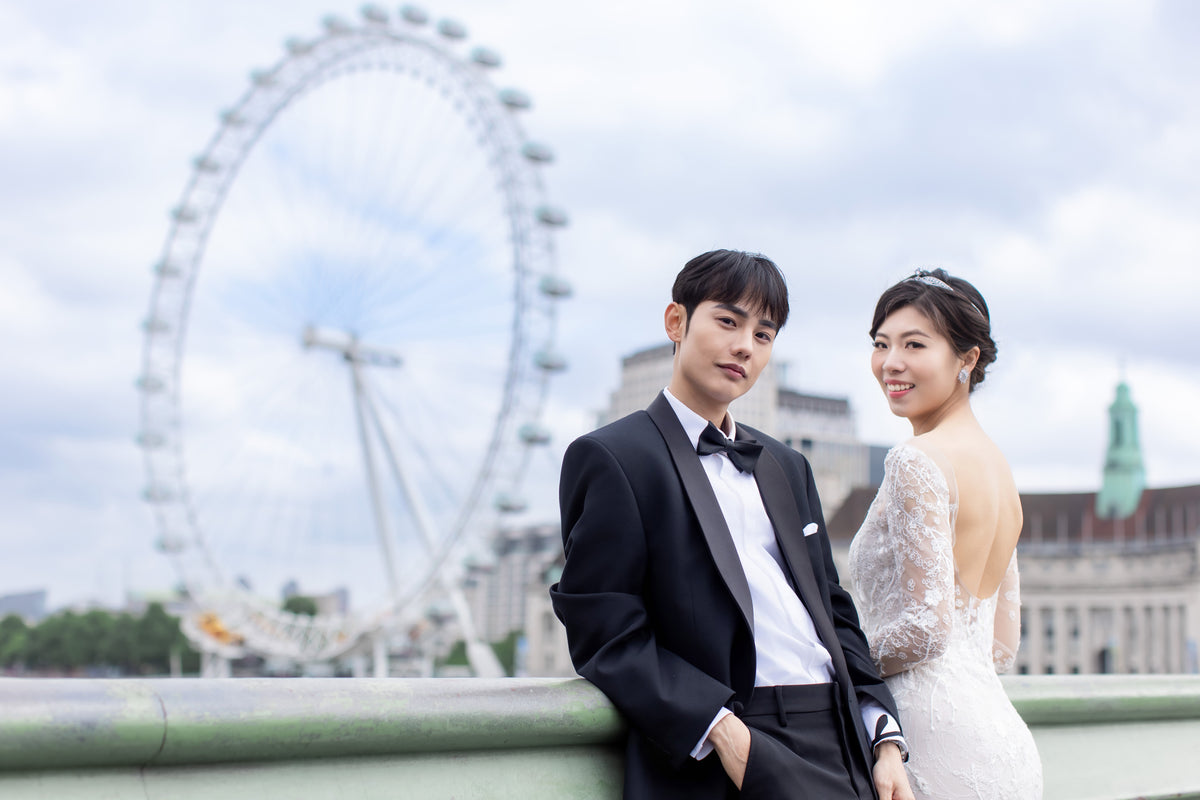 A bride in a white lace wedding gown and a groom in a black tuxedo with a bow tie pose together outdoors, with a large London Eye and city buildings blurred in the background.