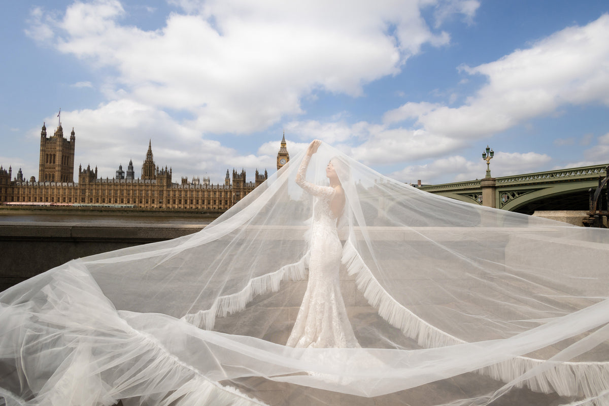 A bride in a white lace wedding gown stands in London with the Houses of Parliament and Westminster Bridge in the background, holding up a long, flowing sheer veil that spreads across the ground under a blue sky with clouds.