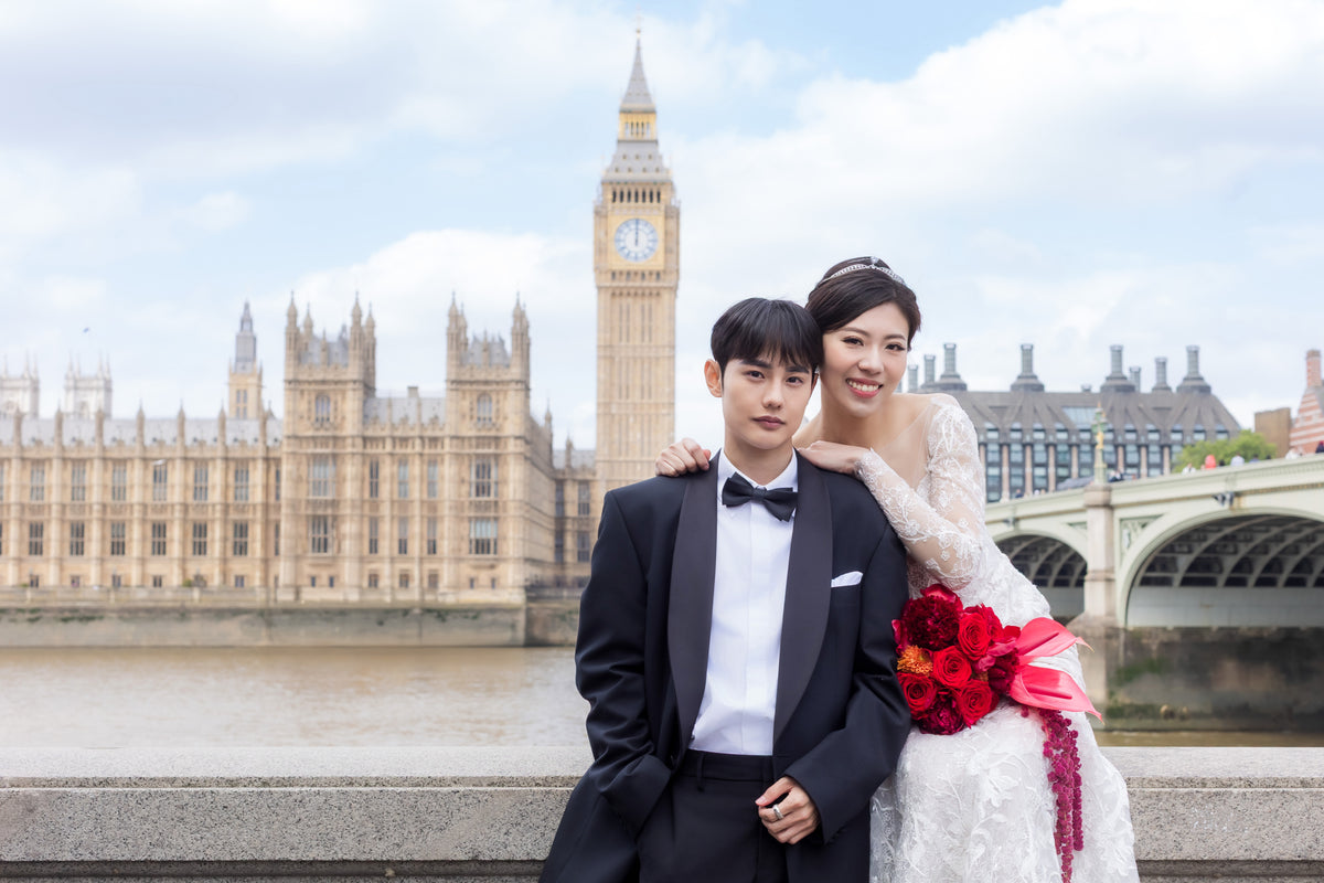 A bride and groom standing outdoors in front of London's Big Ben and Houses of Parliament. The groom wears a black tuxedo with a white shirt and black bow tie, and the bride wears a white lace wedding gown and holds a red flower bouquet.