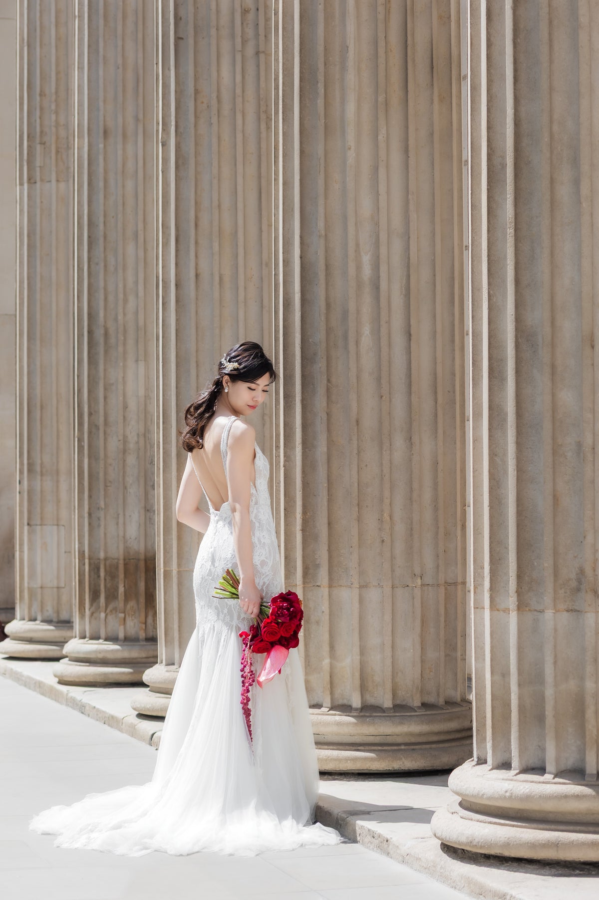 A woman in a white wedding gown stands beside large stone columns, holding a red bouquet.