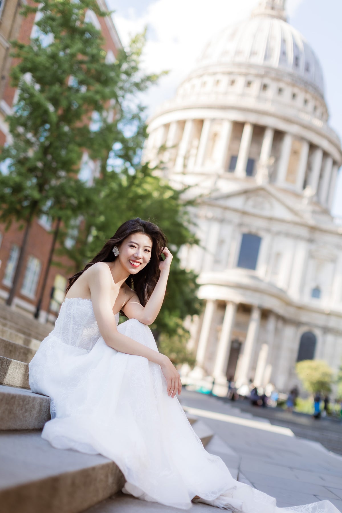 A woman in a white strapless gown sits on stone steps outdoors, with a large domed classical building in the background.