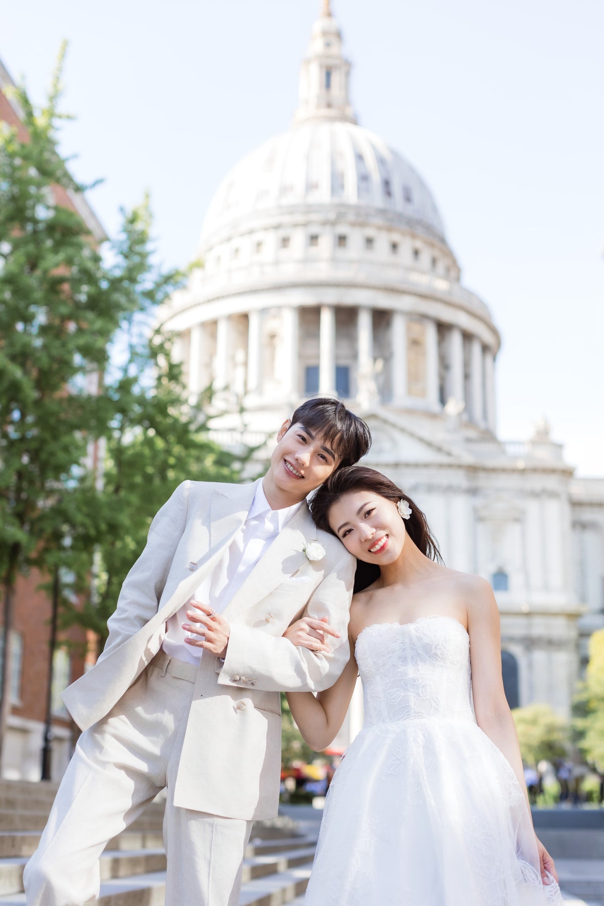 A couple posing in front of a large domed historic building. The man wears a light beige suit with a white shirt and tie, and the woman wears a white strapless gown.