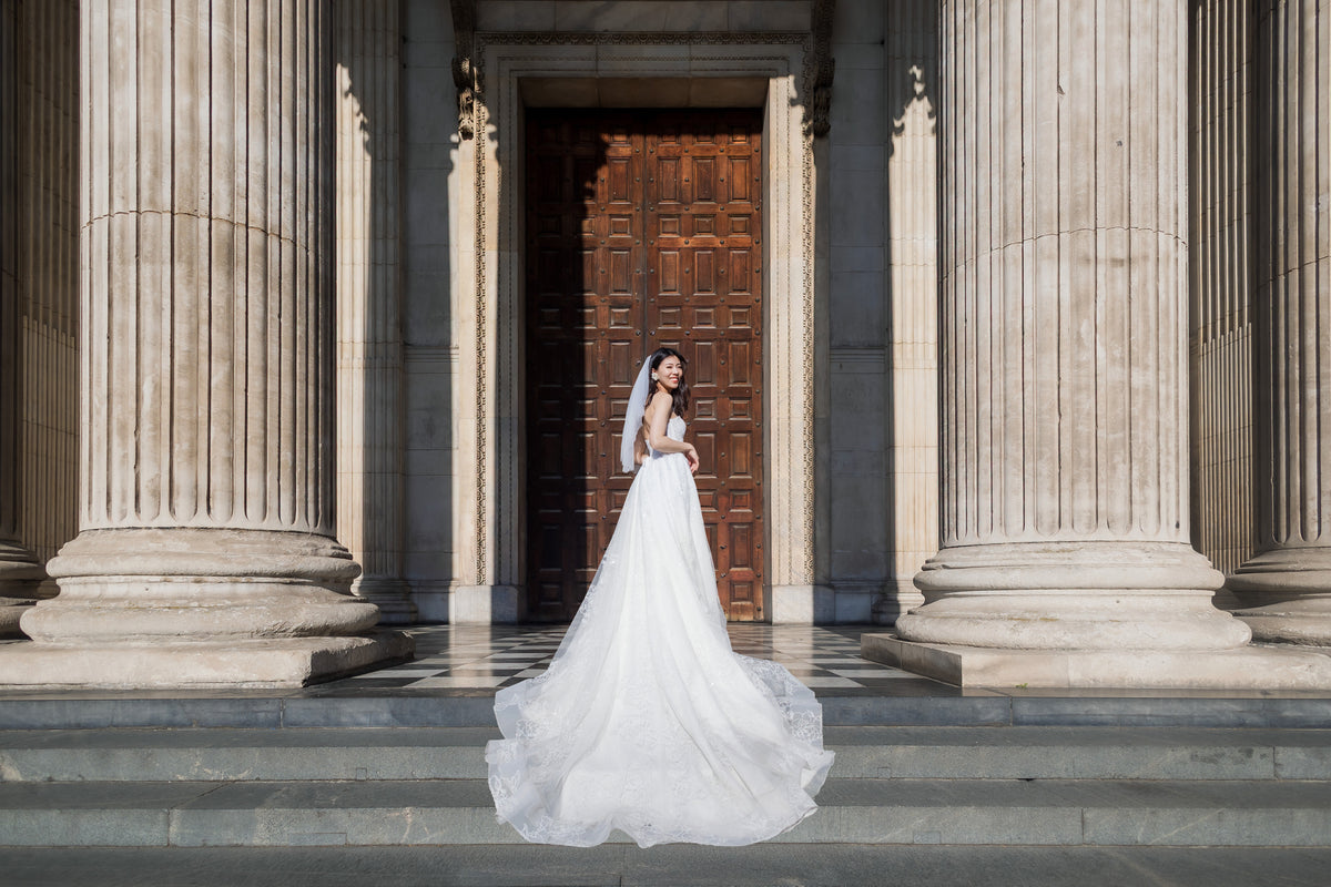A bride in a white wedding gown and veil stands on stone steps in front of a grand classical building with large columns and a dark wooden door.